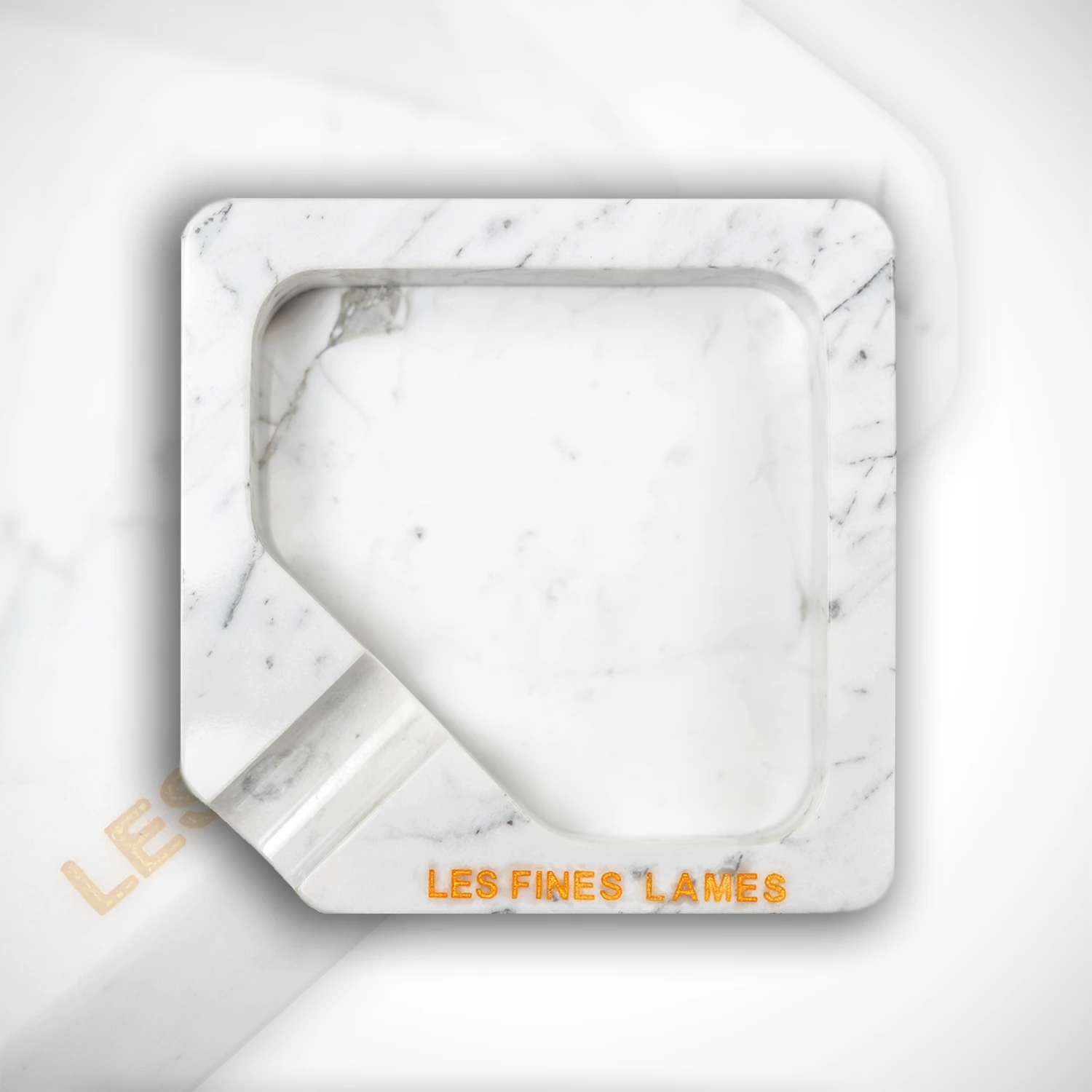 A square white marble ashtray with a recessed center and a small angled notch on the bottom left, featuring the brand name 'LES FINES LAMES' in orange lettering on the front, set against a matching white marble background.