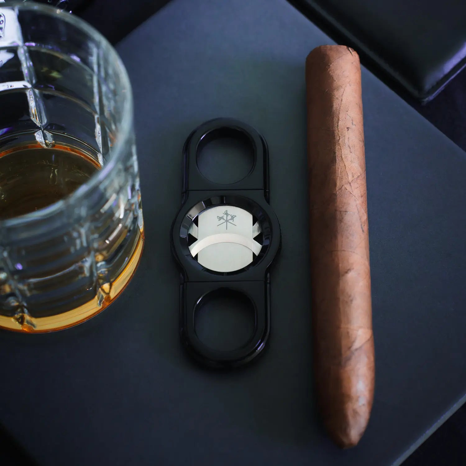La Guillotine - ROTOR Black by LES FINES LAMES, black cigar cutter displayed with a cigar and a glass in a dark setting