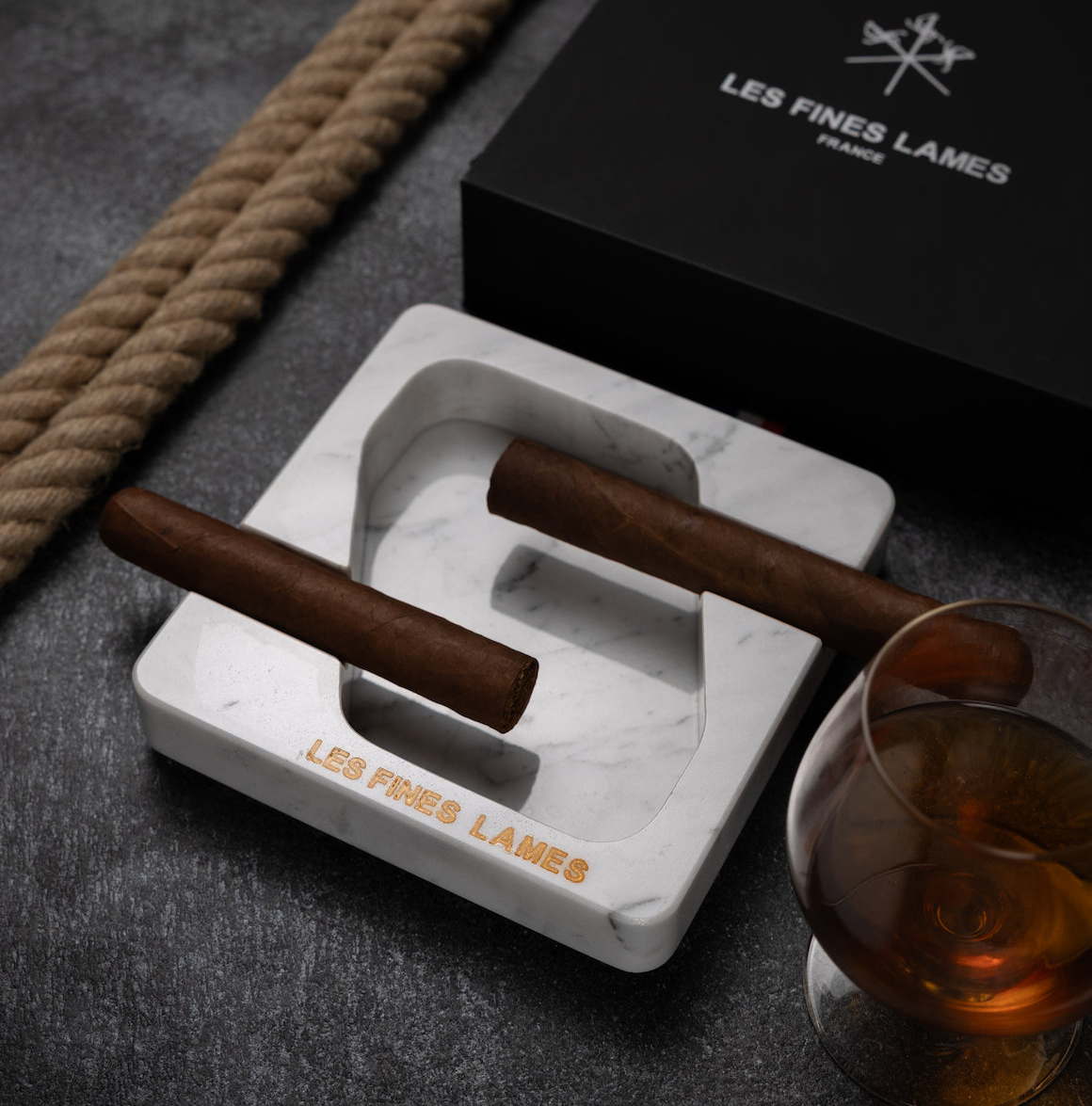 A white marble cigar ashtray engraved with 'LES FINES LAMES FRANCE' sits on a dark gray surface, holding two cigars. A black branded box, a glass of amber liquor, and a rope are styled around it.