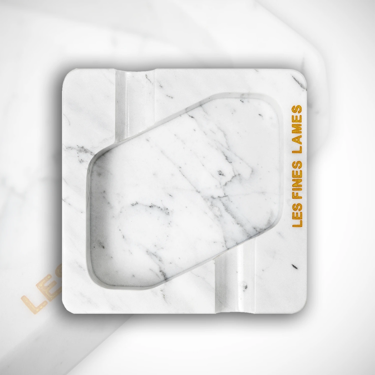 A square white marble-patterned ashtray with a recessed center, featuring the brand name 'LES FINES LAMES' printed in gold on the right side, set against a matching marble background.