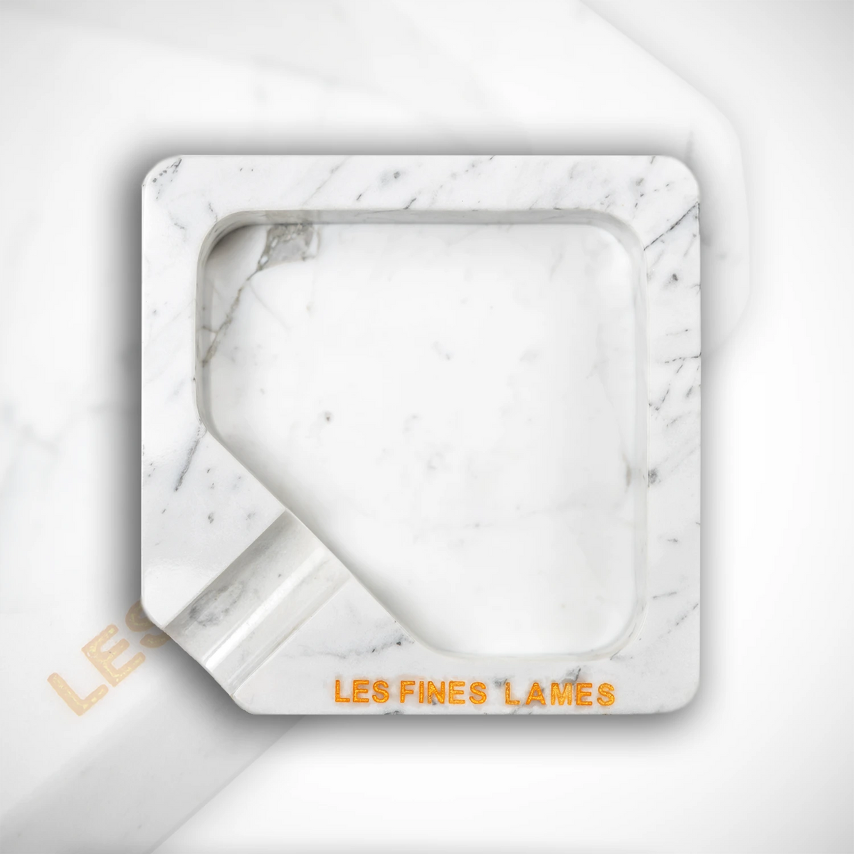 A square white marble ashtray with a recessed center and a small angled notch on the bottom left, featuring the brand name 'LES FINES LAMES' in orange lettering on the front, set against a matching white marble background.
