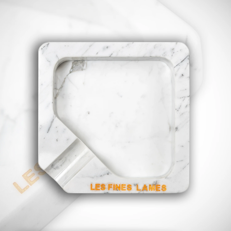 A square white marble ashtray with a recessed center and a small angled notch on the bottom left, featuring the brand name 'LES FINES LAMES' in orange lettering on the front, set against a matching white marble background.