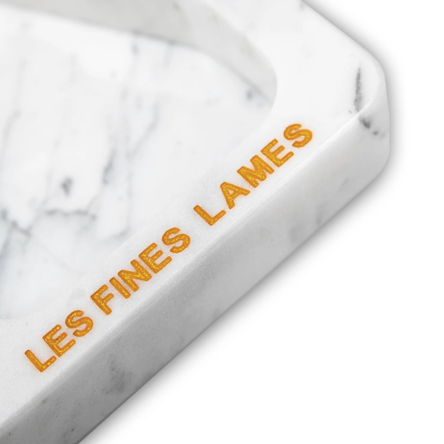 A close-up of a white marble tray corner with the words 'LES FINES LAMES' engraved in gold lettering, set against a clean white background.