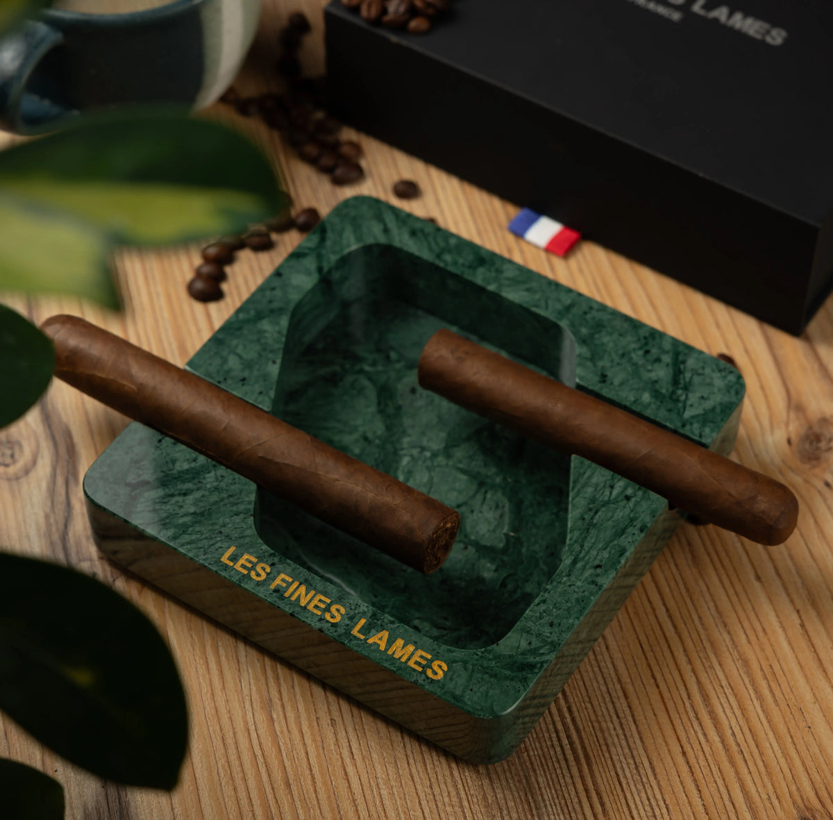 A green marbled square ashtray with gold 'LES FINES LAMES' branding, holding two cigars on a wooden tabletop with coffee beans and a black branded box in the background.