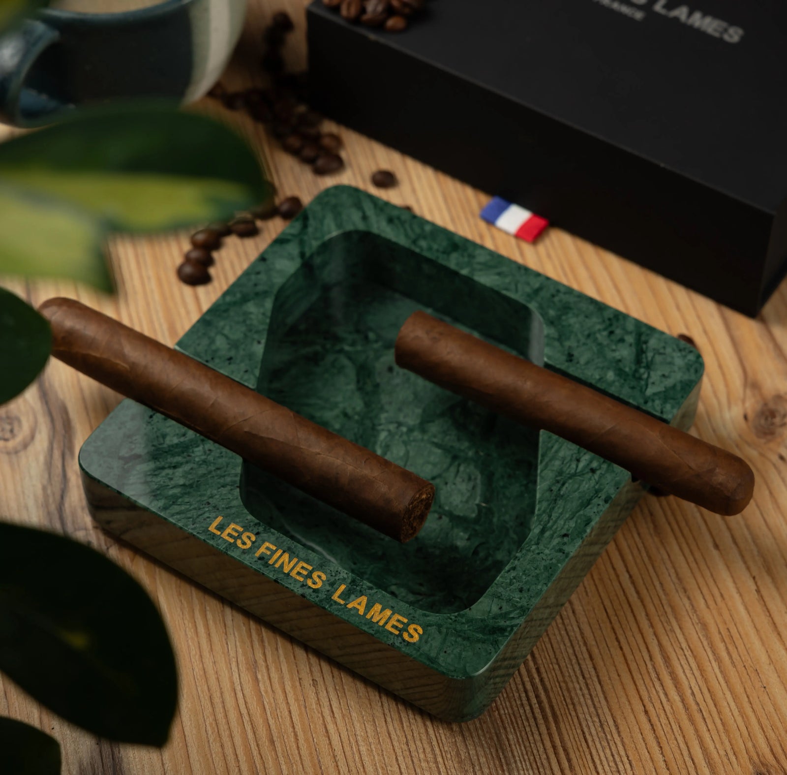A green marbled square ashtray with gold 'LES FINES LAMES' branding, holding two cigars on a wooden tabletop with coffee beans and a black branded box in the background.