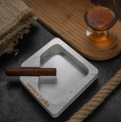 A square white marble ashtray with a cigar resting across one corner, styled on a dark textured surface with a wooden tray, a glass of amber liquor, burlap, and rope nearby. The ashtray is engraved with 'LES FINES LAMES'.